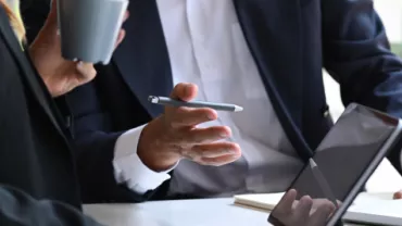 Foco nas mãos de duas pessoas. Uma delas está digitando no computador e a outra está segurando um copo de café, dando a entender que estão em um momento produtivo de trabalho. 