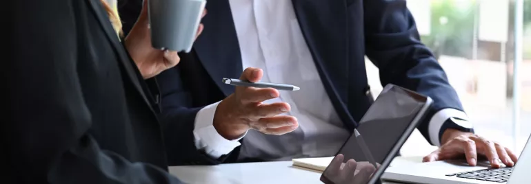 Foco nas mãos de duas pessoas. Uma delas está digitando no computador e a outra está segurando um copo de café, dando a entender que estão em um momento produtivo de trabalho. 